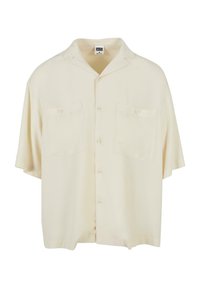 Chemise à manches courtes, ample, en tissu crème, avec un col, deux poches avant et une fermeture à boutons sur le devant. Texture lisse, design minimal.
