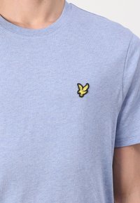 T-shirt en coton bleu clair à manches courtes et col rond. Présente un petit logo d'oiseau noir et jaune brodé sur la poitrine.