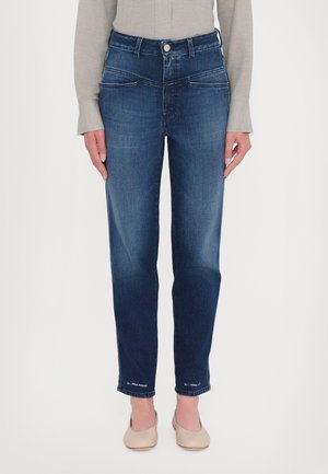 Femme portant un jean slim bleu foncé et des chaussures beiges, debout devant un fond blanc uni.