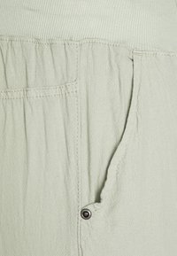 Tissu vert clair ; texture douce avec un léger froissement ; présente une taille côtelée et une poche latérale avec un bouton-pression noir.