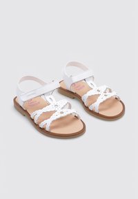 Sandalias blancas con tiras trenzadas y un diseño de corte floral, correa de tobillo ajustable y suela en color marrón. Adecuadas para niños.
