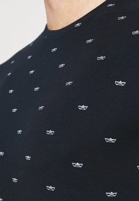 Marineblaues T-Shirt aus Baumwolle, mit einem sich wiederholenden Muster aus kleinen weißen Papierbooten. Rundhalsausschnitt und kurze Ärmel. Glatte Textur.