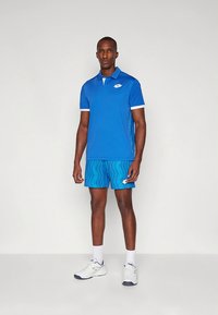 Blå pikétröja med korta ärmar, vit krage och logo, ihop med mönstrade blå shorts. Bärs med vita sportskor och strumpor.