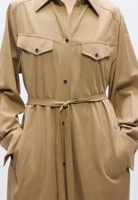 Μπεζ πουκαμισοφορεμα με δύο τσέπες στο στήθος, ζώνη στη μέση και μακριά μανίκια. Διαθέτει μαύρα κουμπιά και λεία υφή υφάσματος.