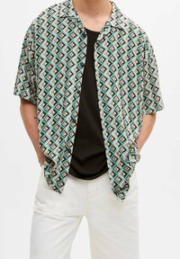 Chemise à manches courtes avec boutonnière, présentant un motif en losanges bleu, vert, orange et noir, réalisée en tissu léger, portée par-dessus un haut noir.