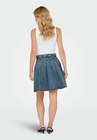 Jupe en denim à design plissé, de couleur bleu foncé, avec une taille. Le modèle la porte avec un débardeur blanc et des sandales à talons noires.