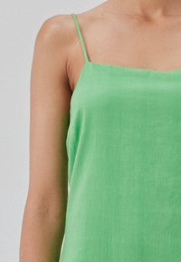 Top sans manches vert en tissu léger. Il dispose de fines bretelles et d'une texture lisse, avec un design simple et minimal.