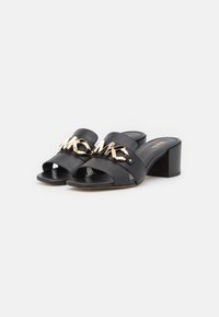 Black leather heeled sandals with open toe, featuring a gold metal chain detail across the upper. Square heel with a smooth texture.