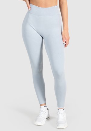 Frau trägt hellblaue Leggings mit hoher Taille und weiße Sneakers und steht vor einem einfarbigen Hintergrund.