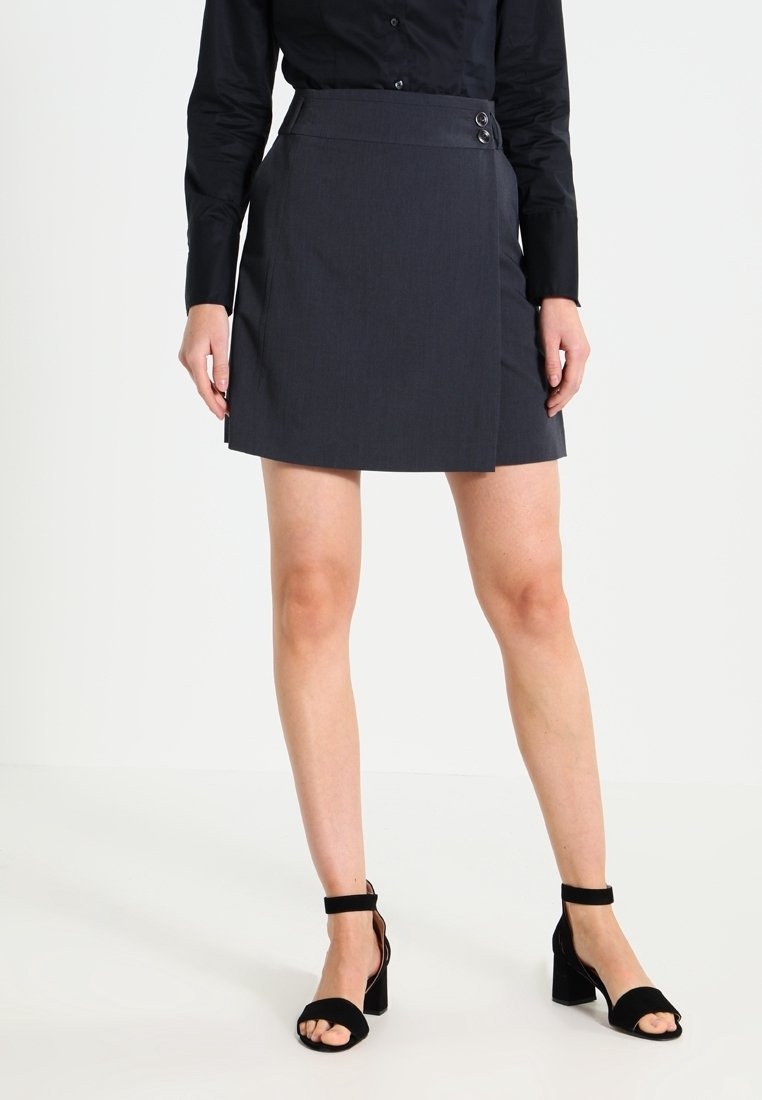 Femme portant une chemise noire boutonnée, une mini-jupe gris foncé avec des boutons sur le côté et des sandales à talons carrés avec bride à la cheville, debout sur un sol blanc.