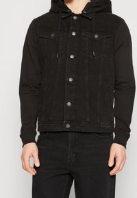 Svart denimjacka med huva, utrustad med knäppningar, bröstfickor och en kontrasterande tygtextur från ärmarna.