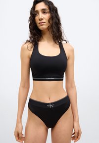 Γυναίκα με σγουρά μαλλιά που φοράει μαύρο αθλητικό σουτιέν Calvin Klein και ταιριαστό εσώρουχο, όρθια μπροστά σε απλό λευκό φόντο.