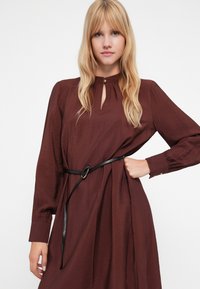 Μακριά μπορντό φόρεμα με ντεκολτέ τύπου κλειδώματος, μακριά μανίκια και μαύρη ζώνη με κυκλικό κούμπωμα. Υφασμάτινη επιφάνεια με λεπτομέρεια συγκέντρωσης στη μέση.