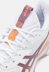 Hvit sportsko med mesh-overdel, oransje og lilla detaljer, og et distinktivt sidemerke. Har skolisser og en teksturert tunge.