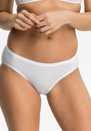Woman wearing white cotton underwear and a matching bra, hands clasped near her waist, against a plain light background.