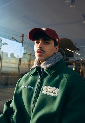 Mladý muž s fúzmi, ktorý má na hlave bordovú šiltovku a oblečenú zelenú bundu s kožušinovým golierom, sedí vo vnútri blízko odrazivého okna s stoličkami za ním.