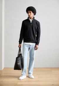 Black zip-neck sweatshirt, light blue straight-leg jeans, white sneakers, holding a black tote bag, with a textured surface and minimal design.