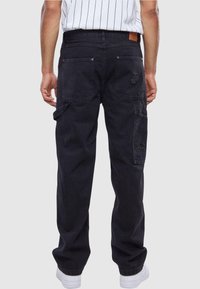 Pantaloni cargo neri con molte tasche, design a gamba dritta e tessuto testurizzato. Presentano una toppa logo sul retro e sneakers bianche.