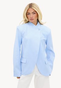 Femme blonde portant un blazer asymétrique bleu clair boutonné et un pantalon blanc, regardant droit devant avec une expression neutre.