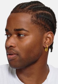 Orecchino a cerchio dorato con un design intrecciato, indossato da una persona con capelli corti e treccia. Il tono della pelle è marrone medio; indossa una camicia bianca.