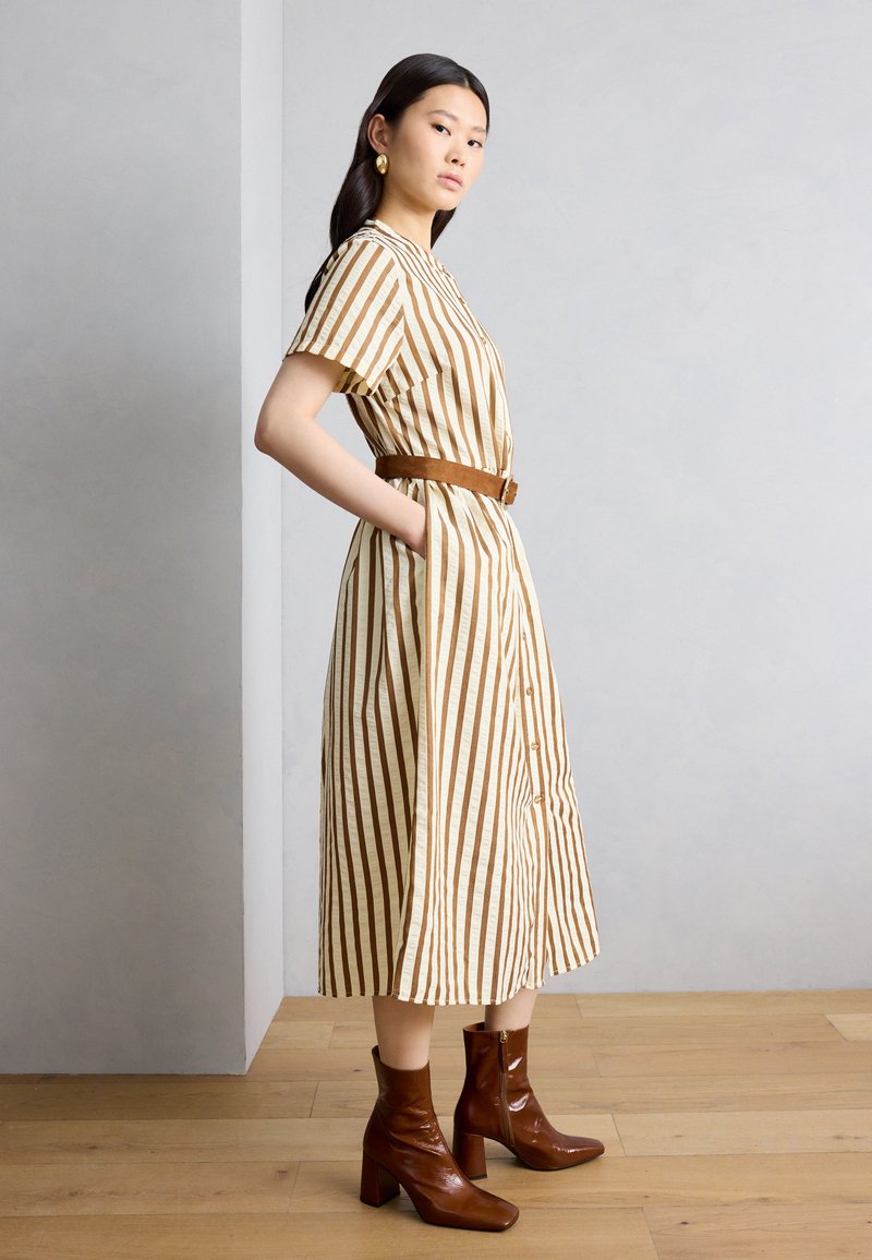 Γυναίκα φορώντας μίντι φόρεμα με κάθετες καφέ και μπεζ ρίγες, με κοντά μανίκια και καφέ ζώνη, συνδυασμένο με καφέ μποτάκια αστραγάλου, στέκεται πλάγια.