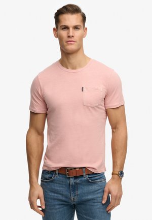 Homme portant un T-shirt rose clair à manches courtes avec poche, jean bleu, ceinture marron et montre-bracelet, debout devant un fond blanc.