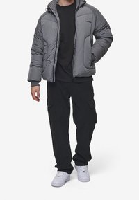 Uomo che indossa un piumino grigio sopra una maglietta nera, pantaloni cargo neri e sneakers bianche, in piedi con una mano in tasca.