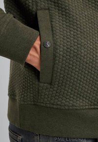 Oliwkowy sweter z teksturowanej tkaniny w splot miodu, z prążkowanymi mankietami oraz boczną kieszenią z czarnym guzikiem.