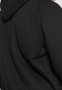 Gros plan sur le tissu d'un sweat à capuche noir montrant la couture de l'épaule et une partie de la capuche attachée sur un fond uni.