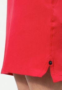 Falda de tela roja con una apariencia texturizada, que presenta un detalle de botón negro y un dobladillo cosido en el lateral.