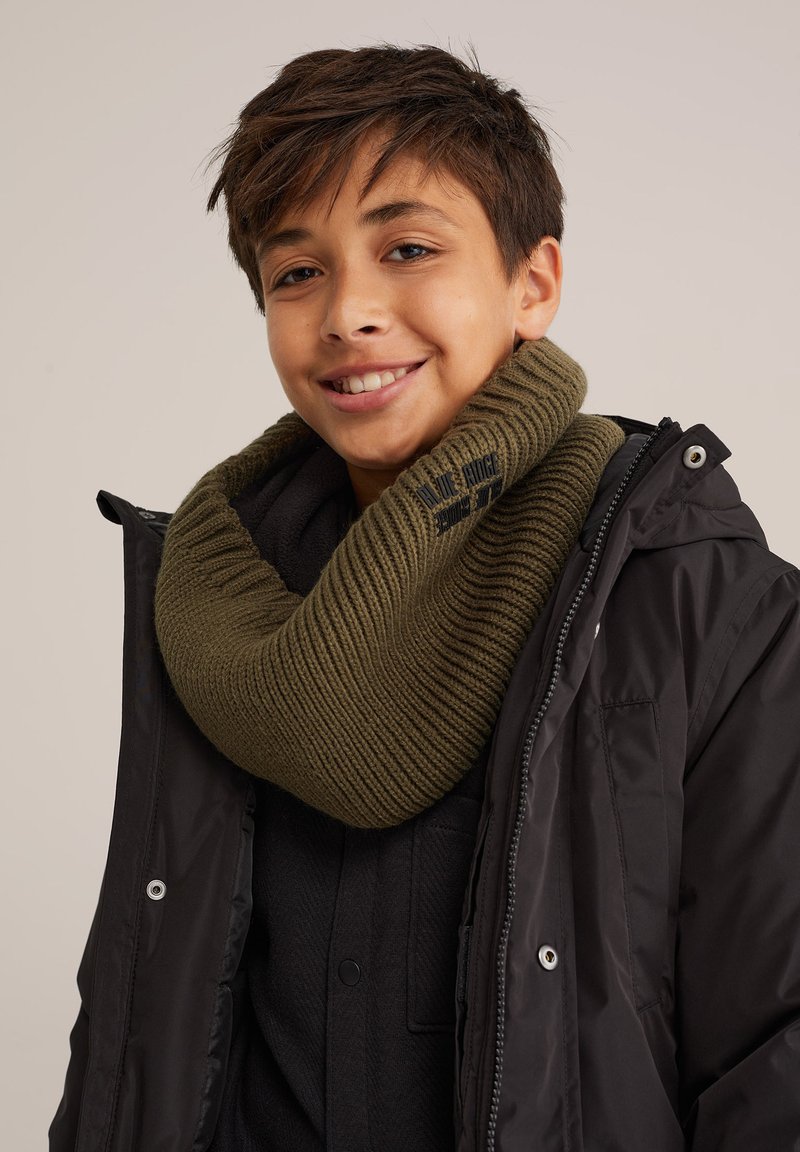 Adīta olīvzaļa kakla šalle ar ribotu tekstūru, kas valkāta virs melna mēteļa. Tai ir logotipa detaļa un plaša forma, kas krīt ap kaklu.