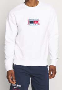Vit sweatshirt i bomullsmaterial med en logotyp i röd, blå och svart i mitten. Ribbad ärm och nederkant, avslappnad passform.