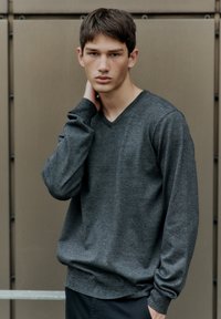 Donkergrijs, gestructureerd gebreid sweater met een V-hals, lange mouwen en geribbelde manchetten. Een relaxte pasvorm, gedragen door een mannelijke model tegen een neutrale achtergrond.