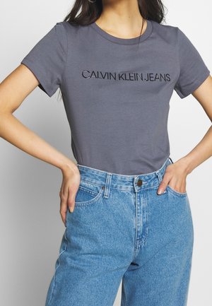 Gray cotton T-shirt featuring "CALVIN KLEIN JEANS" in black lettering, paired with high-waisted light blue denim jeans.