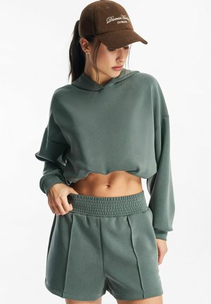 Młoda kobieta w zielonej krótkiej bluzie z kapturem i dopasowanych szortach, poprawiająca pas, nosząca brązową czapkę z daszkiem, patrząca w dół na białym tle.