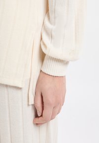 Helles cremefarbenes Oberteil mit vertikal gerippter Textur, ausgestattet mit geripptem Bündchen und seitlichem Schlitzdetail. Hand teilweise sichtbar, ruht auf dem Stoff.