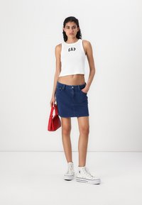 White cropped tank top with "GAP" logo, dark blue denim skirt, white high-top sneakers, and a small red handbag.
