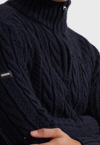 Marinblå kabelstickad tröja med hög krage som har tjocka ribbningar, en frontdragkedja och en strukturerad väv, med en etikettdetalj på vänster ärm.