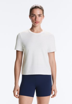 Femme debout, de face, portant un t-shirt blanc à manches courtes avec de subtiles rayures horizontales et un short bleu marine.