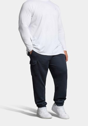 Persoon draagt een wit shirt met lange mouwen, zwarte cargo joggerbroek en witte sneakers, staat met handen in de zakken tegen een effen achtergrond.