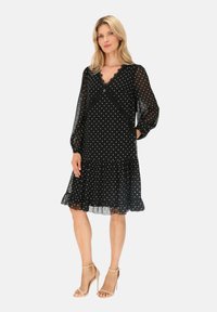 Czarna sukienka w kropki, uszyta z przezroczystego materiału, z długimi rękawami, falbaniastym dołem i dekoltem w kształcie V wykończonym koronką. Połączona z beżowymi sandałami.