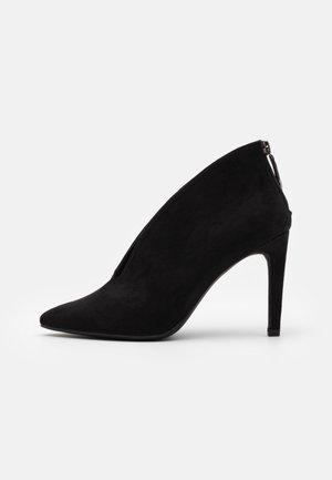 Sort ruskind højhælet sko med spids tå, tynd stiletthæl og lynlås bagpå på en hvid baggrund.