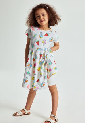Meisje met krullend haar dat een witte jurk draagt bedrukt met kleurrijke ijsjes- en fruitpatronen, witte legging en witte sandalen.