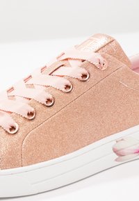 Zapatillas con glitter rosa dorado, cordones rosas, ojales metálicos y detalle de estampado floral en la suela blanca. Textura suave, punta redonda.