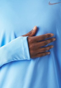 Mörkhyad hand med tumhål i en ljusblå långärmad skjorta, vilande på bröstet nära en liten Nike-logotyp.