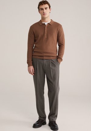 Man draagt een getextureerde bruine lange- mouwen polo trui, geruite geplooide broek en zwarte leren schoenen, staand met één hand in de zak.