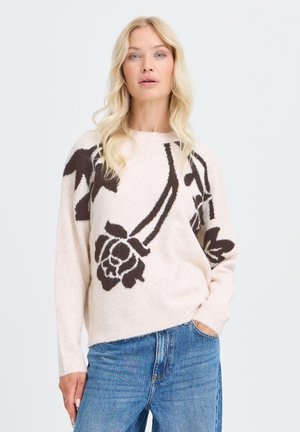 Femme blonde portant un pull beige clair avec de grands motifs floraux foncés et un jean bleu, debout, une main dans la poche.
