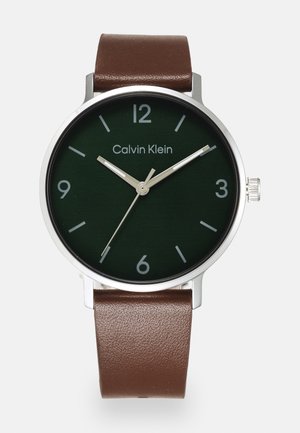 Montre-bracelet Calvin Klein avec cadran vert foncé, boîtier argenté, aiguilles argentées, index gris et bracelet en cuir marron. L'heure affichée est 10:08:36.