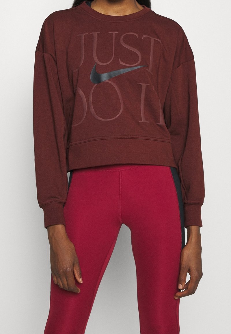 Personne portant un sweat-shirt Nike bordeaux avec le texte "Just Do It" et des leggings bordeaux avec des panneaux latéraux noirs, debout sur un fond uni.