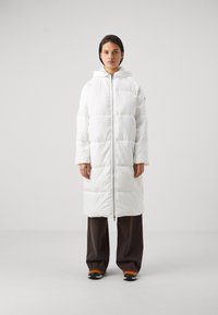 Doudoune blanche avec capuche, dotée d'une fermeture éclair intégrale et de sections matelassées horizontales. Portée par-dessus un pantalon large sombre et des baskets.